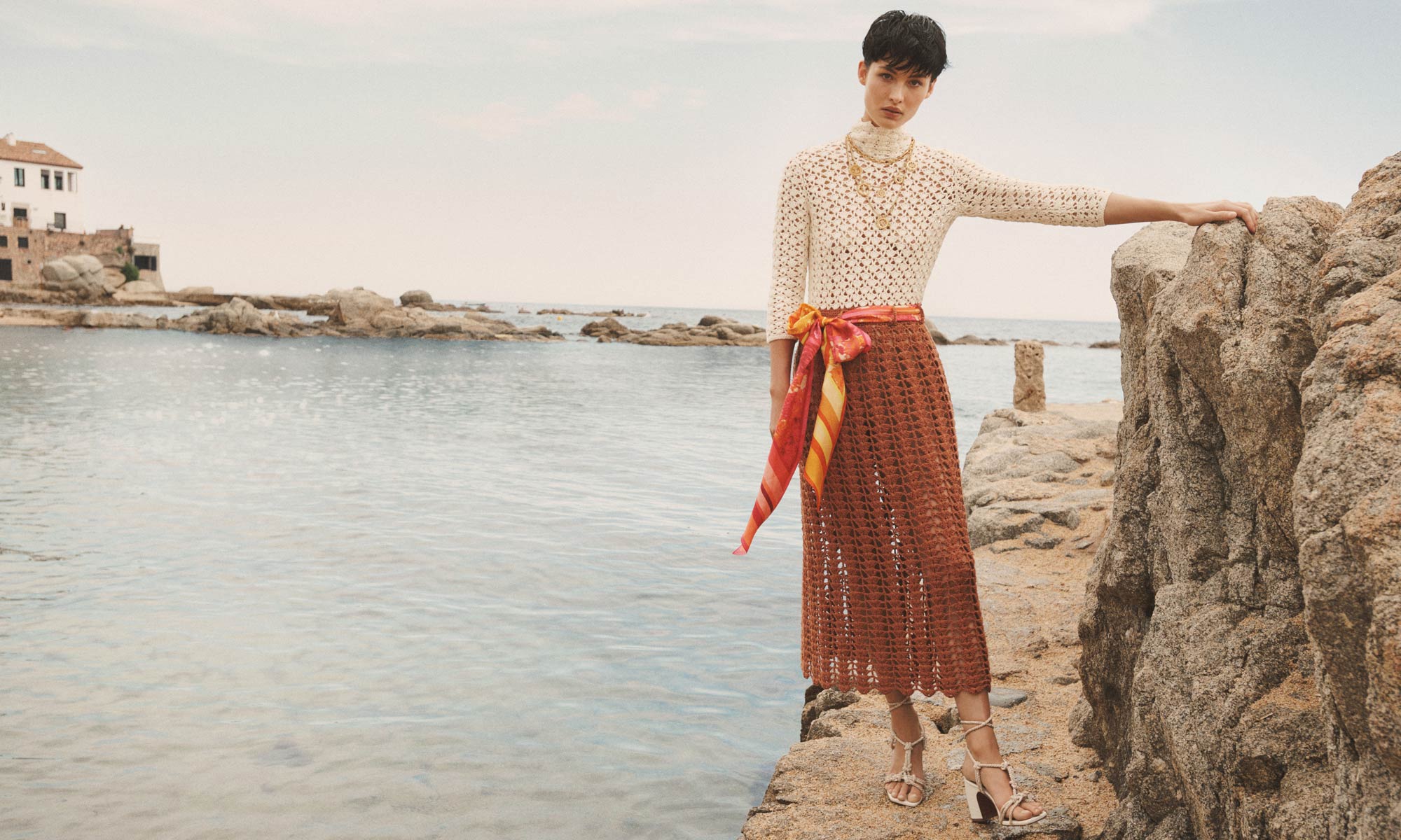 A woman wearing a white and brown crochet dress with a colorful sash poses on rocks by the sea with a building visible in the distance.