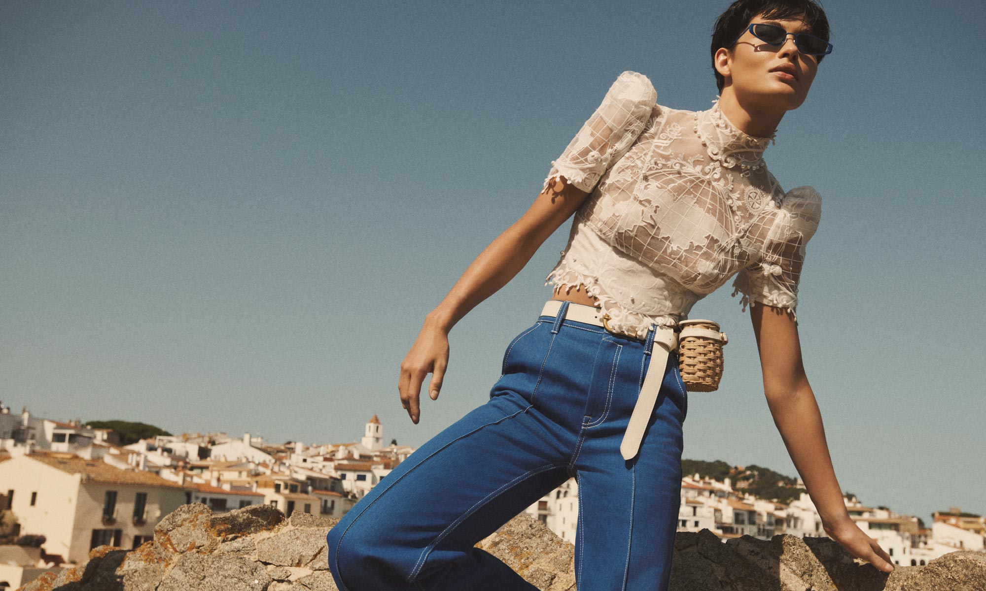 A fashionable woman in a white lacy top and blue jeans stands outdoors against a backdrop of a coastal town and clear blue sky.