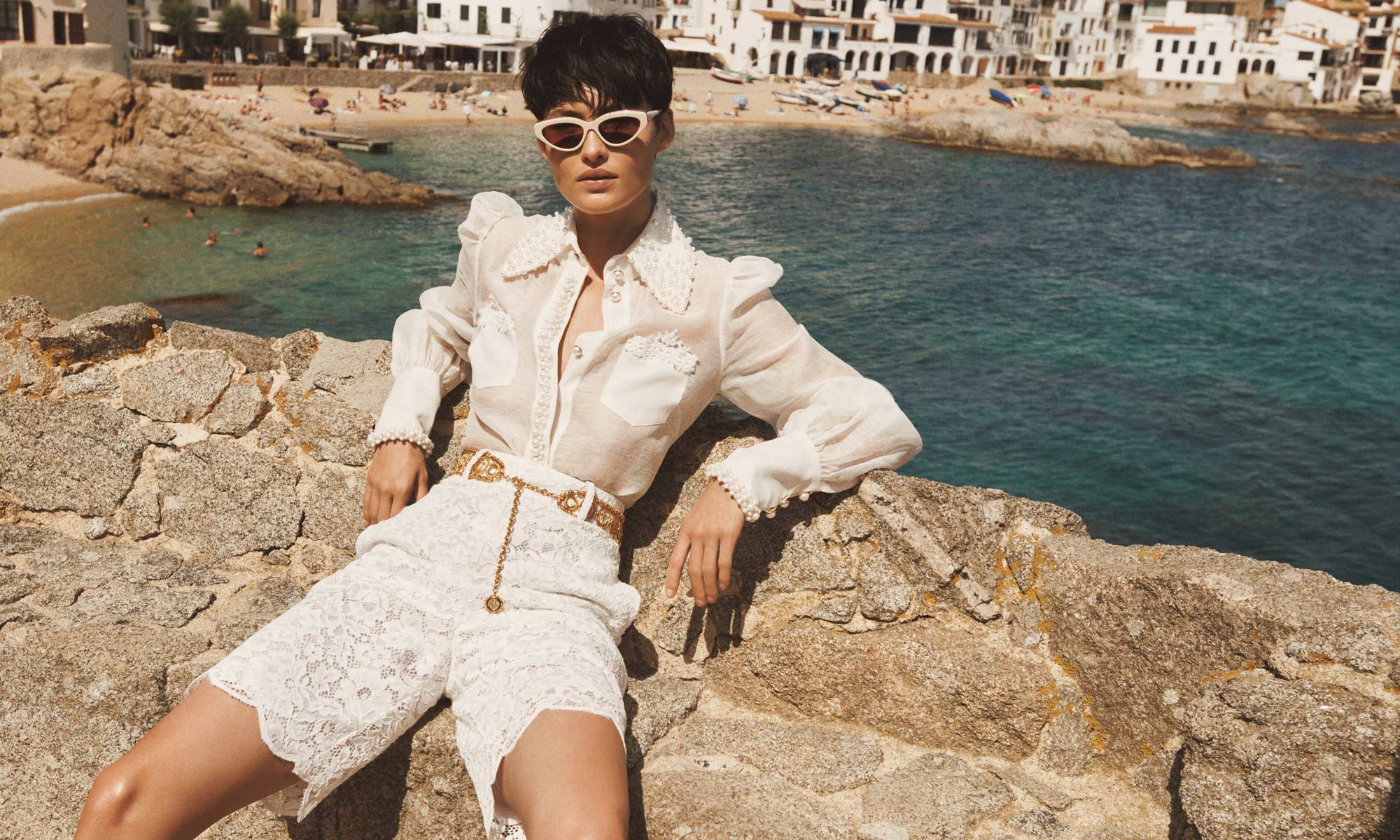 A woman in a white linen blouse and lace shorts poses on a stone wall overlooking the ocean and a beach town with white buildings.