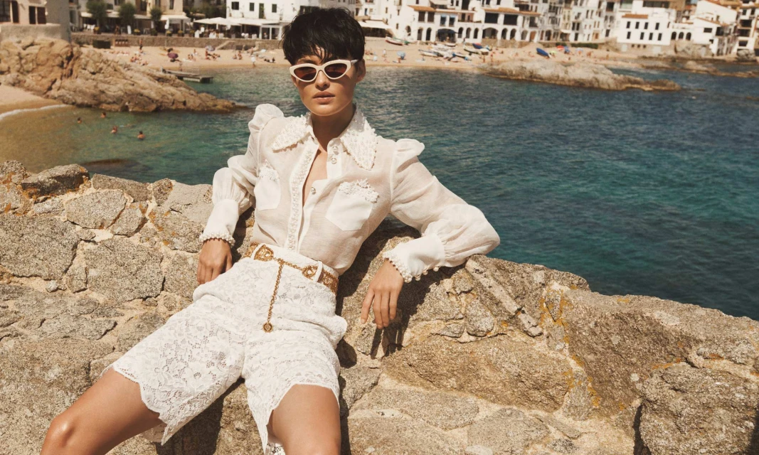 A woman in a white linen blouse and lace shorts poses on a stone wall overlooking the ocean and a beach town with white buildings.