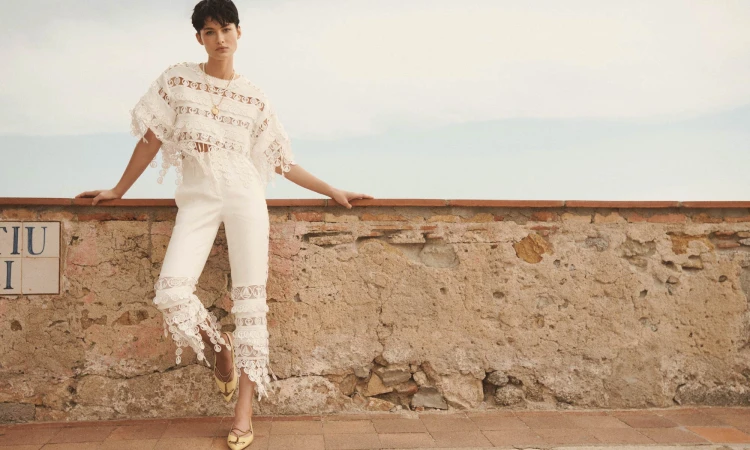 Model wearing a white lace top and trousers posing against a weathered stone wall under an overcast sky for Zimmermann Resort RTW.