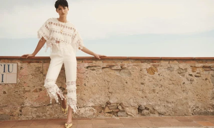 Model wearing a white lace top and trousers posing against a weathered stone wall under an overcast sky for Zimmermann Resort RTW.