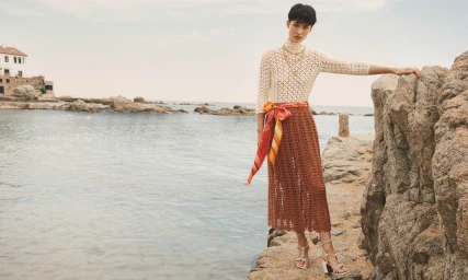 Model standing on a rocky shore by the ocean wearing a white knit top and a patterned brown skirt.