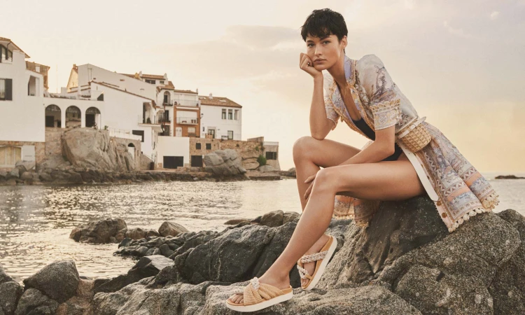 Model with short dark hair wearing a patterned tunic seated on coastal rocks against a Mediterranean town backdrop