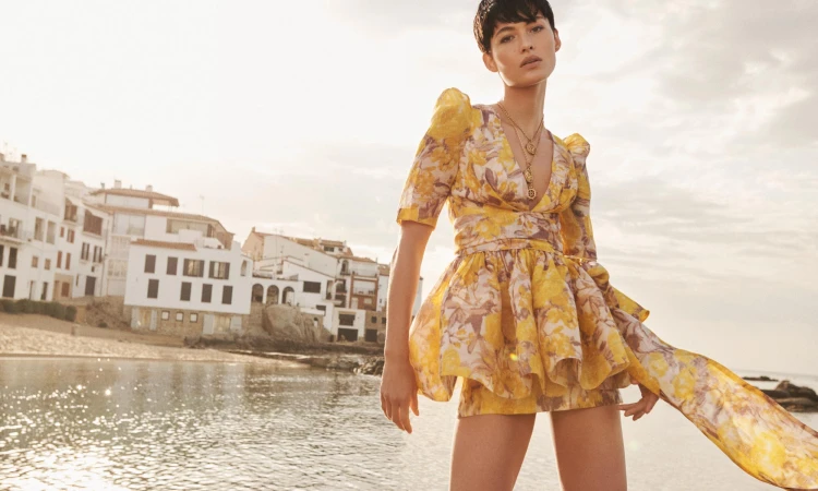 Model in a yellow floral dress standing outdoors near a coastal town and water for Zimmermann Resort RTW.