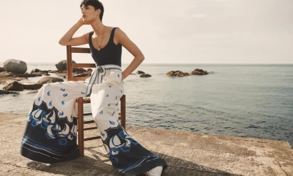 Model with short dark hair seated on a wooden chair wearing a blue top and patterned wide-leg trousers by the coast.
