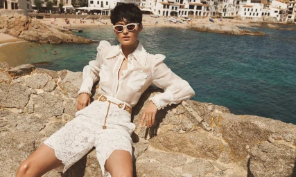 Model wearing a white lace shirt and shorts, sunglasses, and a gold chain belt seated against a stone wall by the ocean