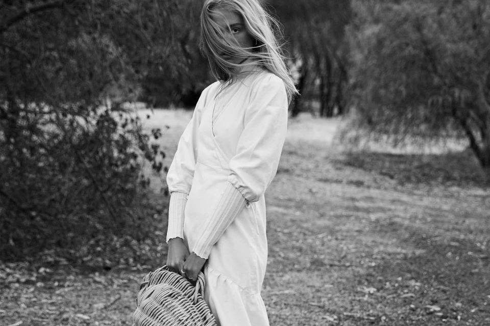 Monochrome portrait of a young woman in a white dress holding a woven basket, standing outdoors in a grassy field surrounded by trees.