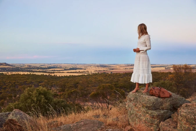 Woman in a white lace dress standing barefoot on a rocky outcrop overlooking a vast landscape under a pale sky