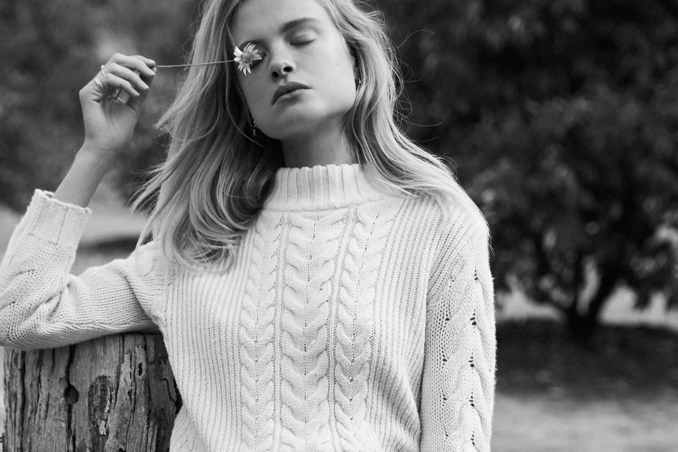 Black and white portrait of a young blonde woman with a daisy flower in front of her eye, wearing a cable knit sweater outdoors.