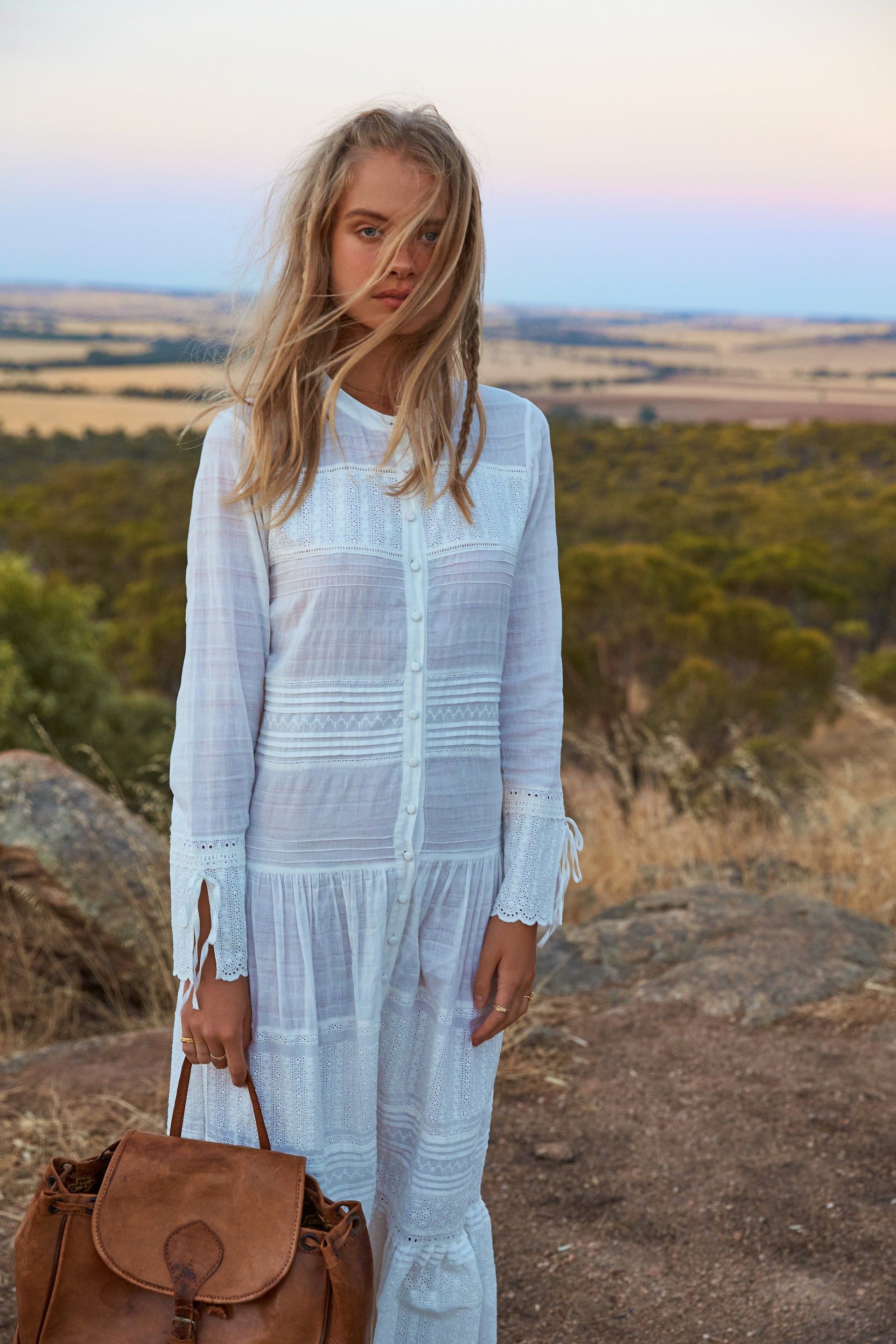Woman in a white dress stands on a hill with a leather backpack. The background is an open field with a sunset sky.