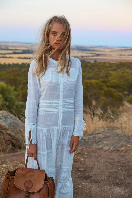 Woman in a white dress stands on a hill with a leather backpack. The background is an open field with a sunset sky.