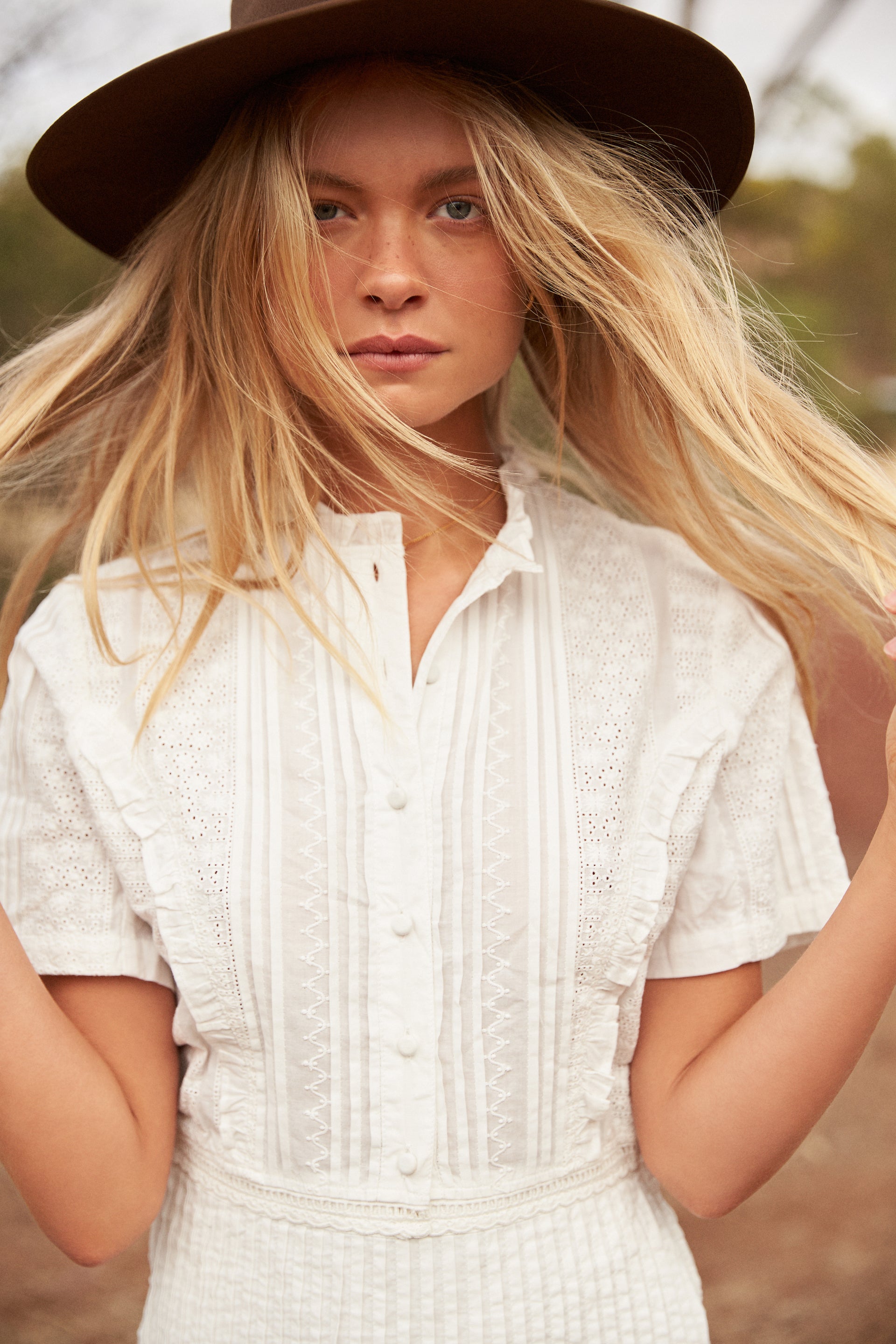 Model wearing a white lace blouse and dark hat with blonde hair blowing in the wind outdoors