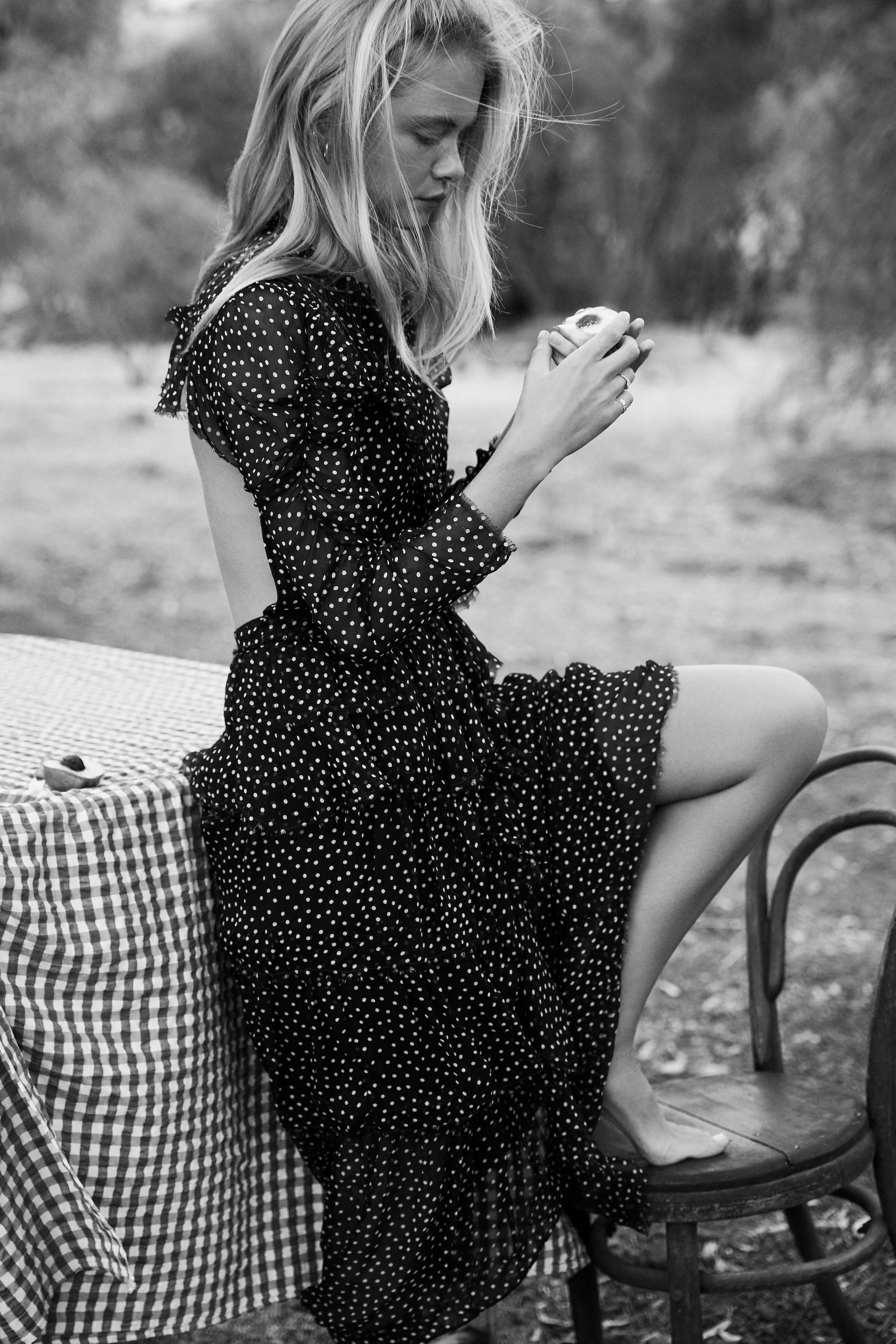 Blonde woman in a polka dot dress sits on a chair next to a gingham-covered table, holding an apple in a black and white outdoor setting.