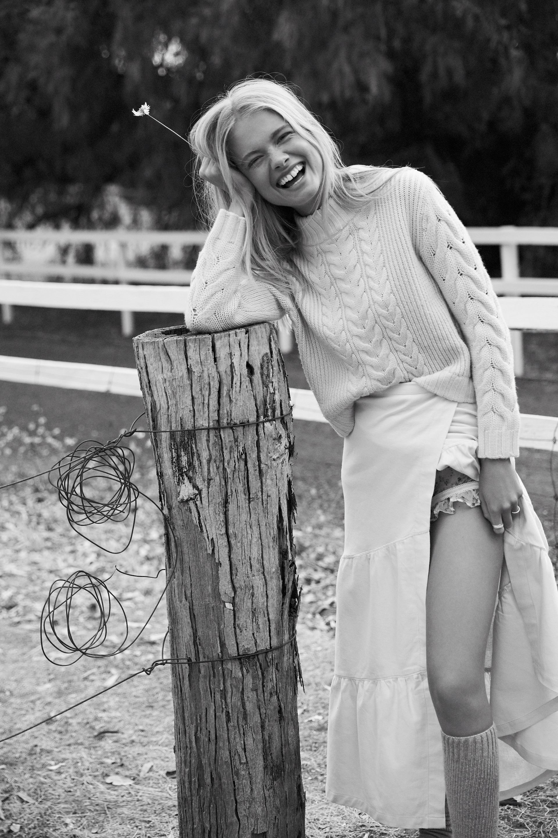 Happy woman smiles leaning on a wooden post, wearing a sweater and a long skirt in a rural setting with a white fence in the background.