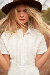 Model wearing a white lace blouse and dark hat with blonde hair blowing in the wind outdoors