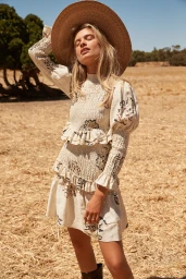 Model in smocked cream dress and large straw hat standing outdoors in dry field under bright sunlight.