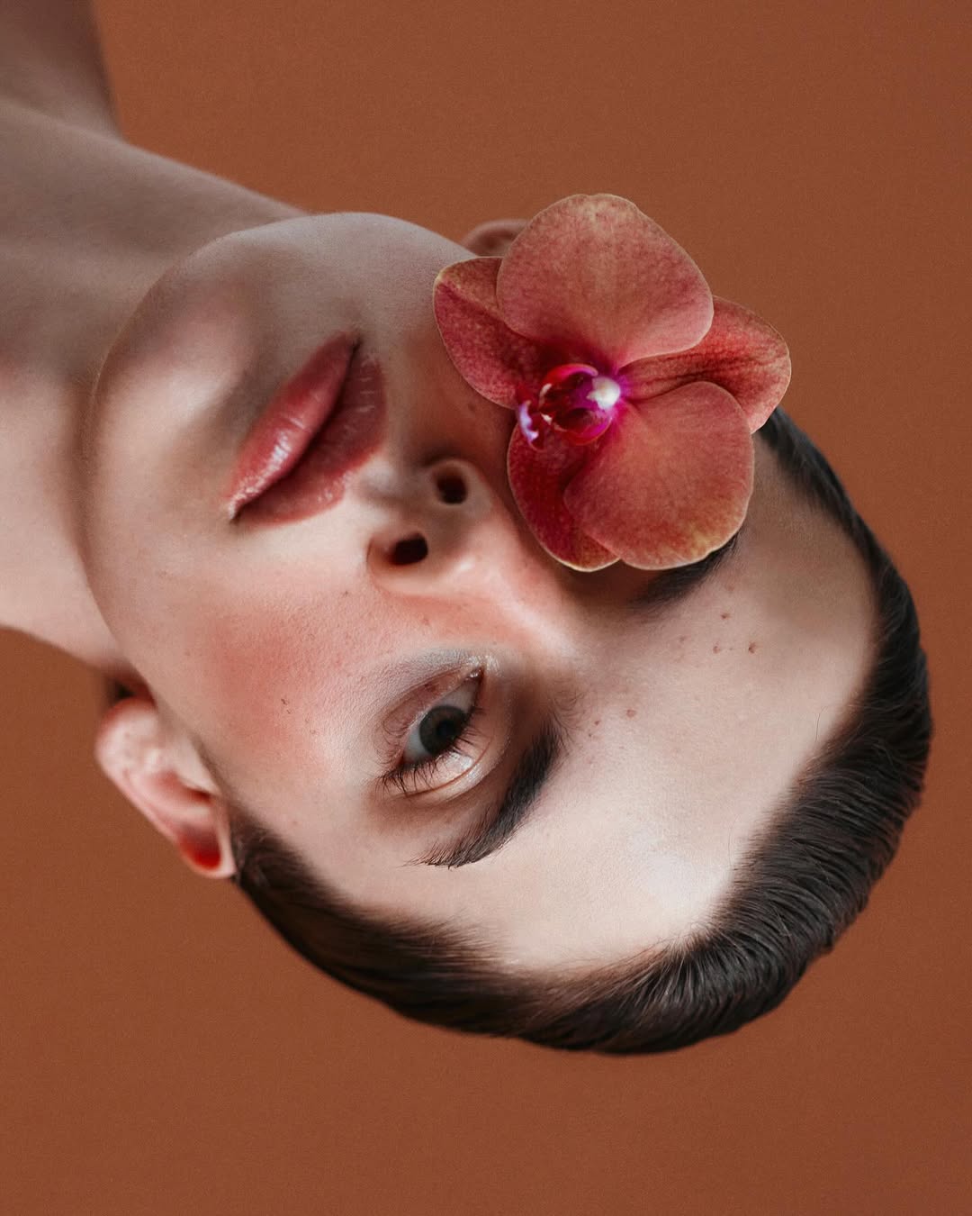 Close-up portrait of a person's face with an orchid flower on their forehead, set against a warm, terracotta-colored background.