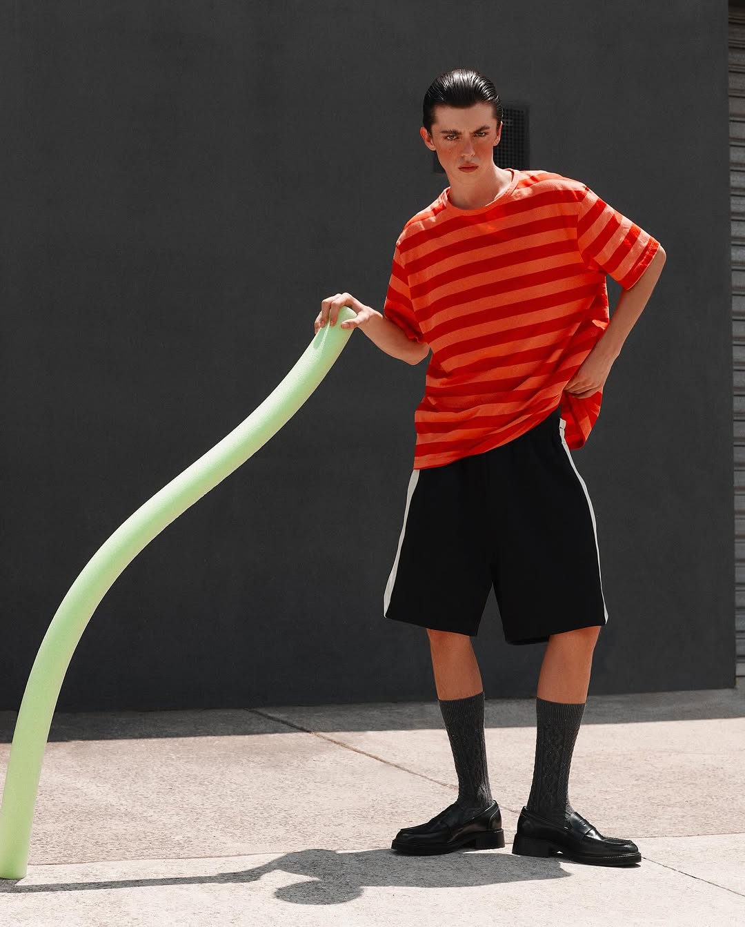 Adult male fashion model poses in an outdoor setting wearing a striped shirt, black shorts, socks, and dress shoes, holding a green balloon.