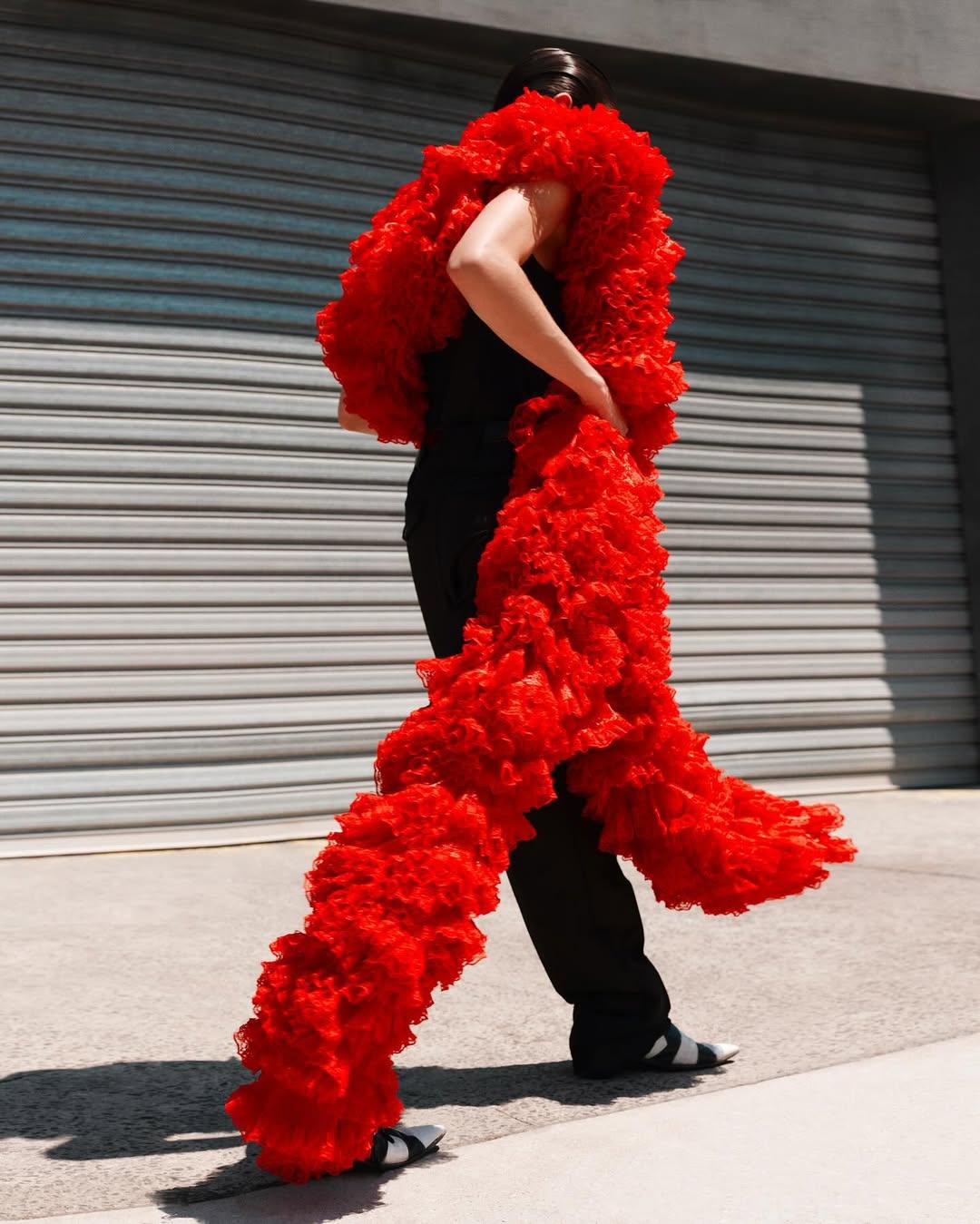 Adult female in black pants and top poses outdoors wearing a long red ruffled scarf, standing near a gray metal garage door in sunlight.