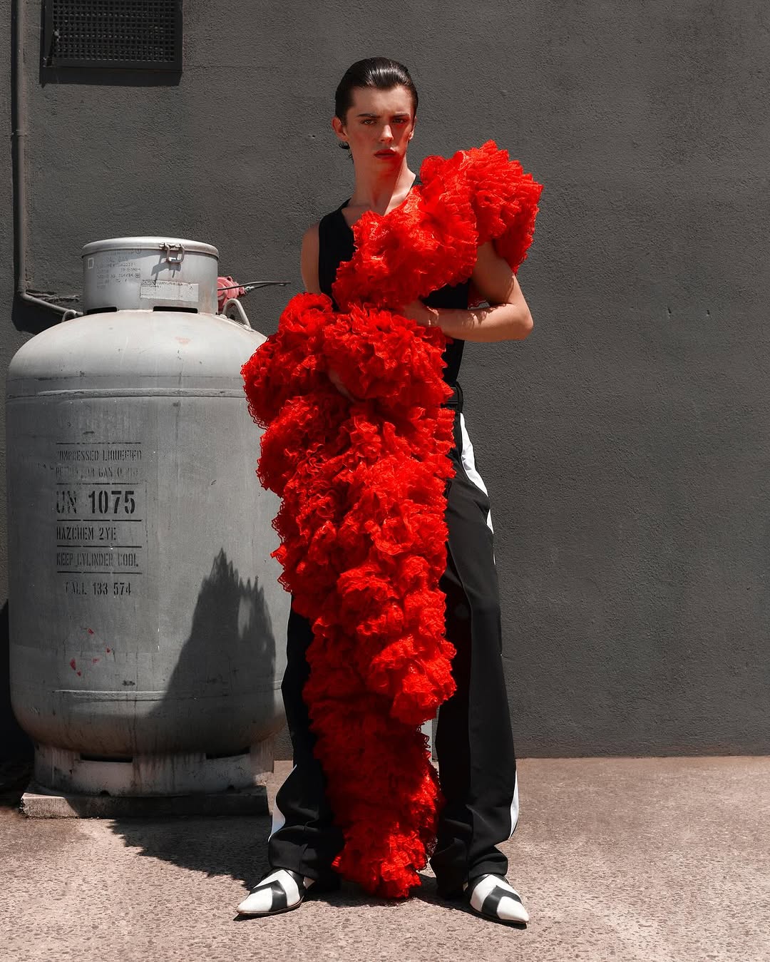 A person standing in front of a grey wall next to a propane tank while wearing a black outfit and holding a large red feathery boa.