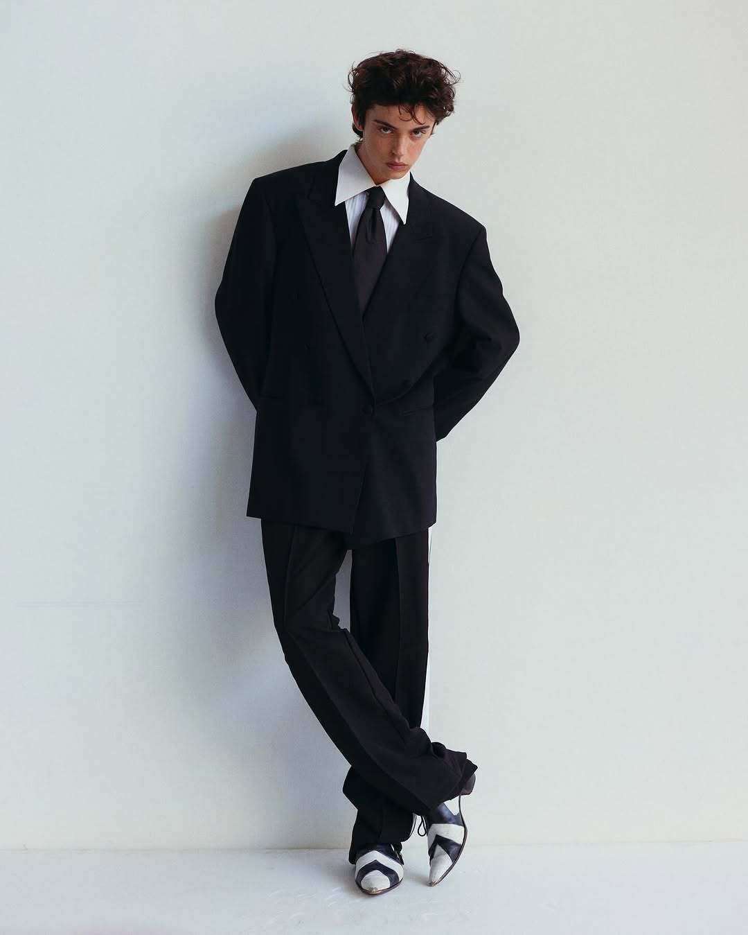 Man in black suit with wide collared white shirt standing in front of a white wall looking at the camera in a studio environment.