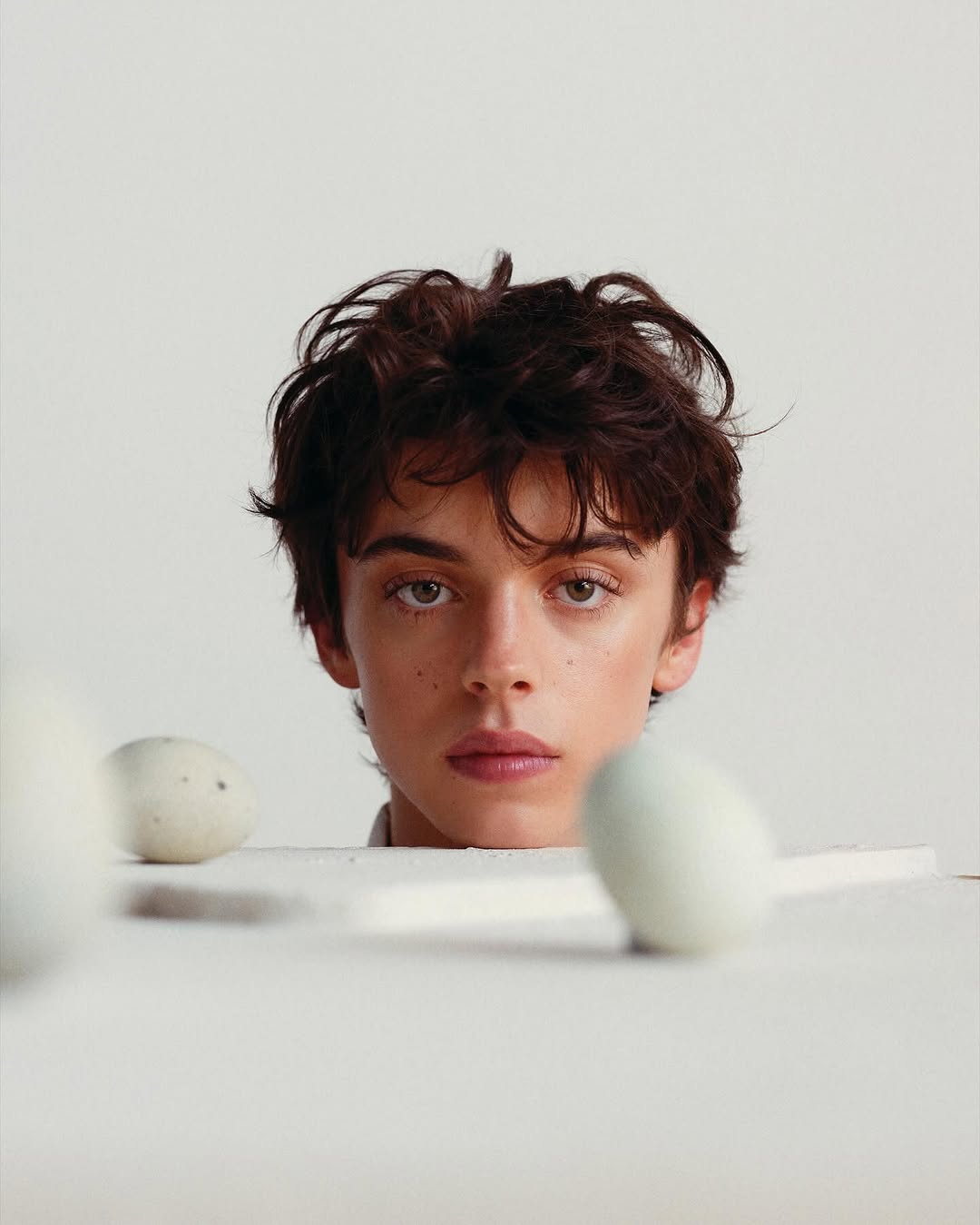 Close-up portrait of a young adult with dark hair, light skin, and light eyes, gazing forward with neutral expression. Eggs in foreground.