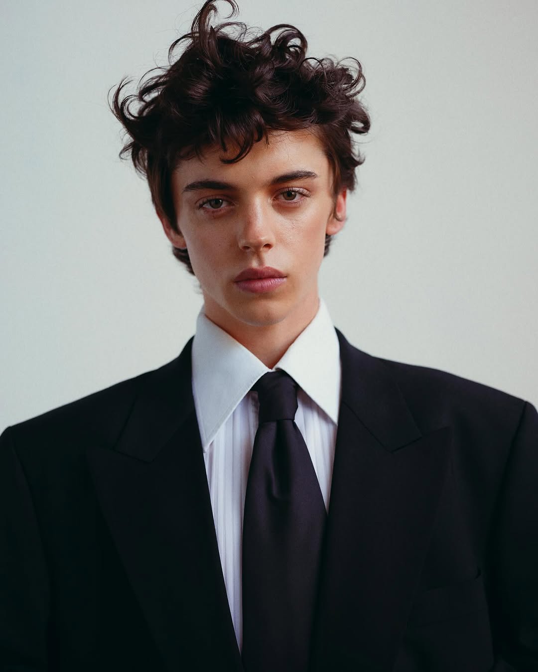 Headshot of a stylish brunette male in a black suit, featuring a white collared shirt and black tie against a light neutral backdrop.