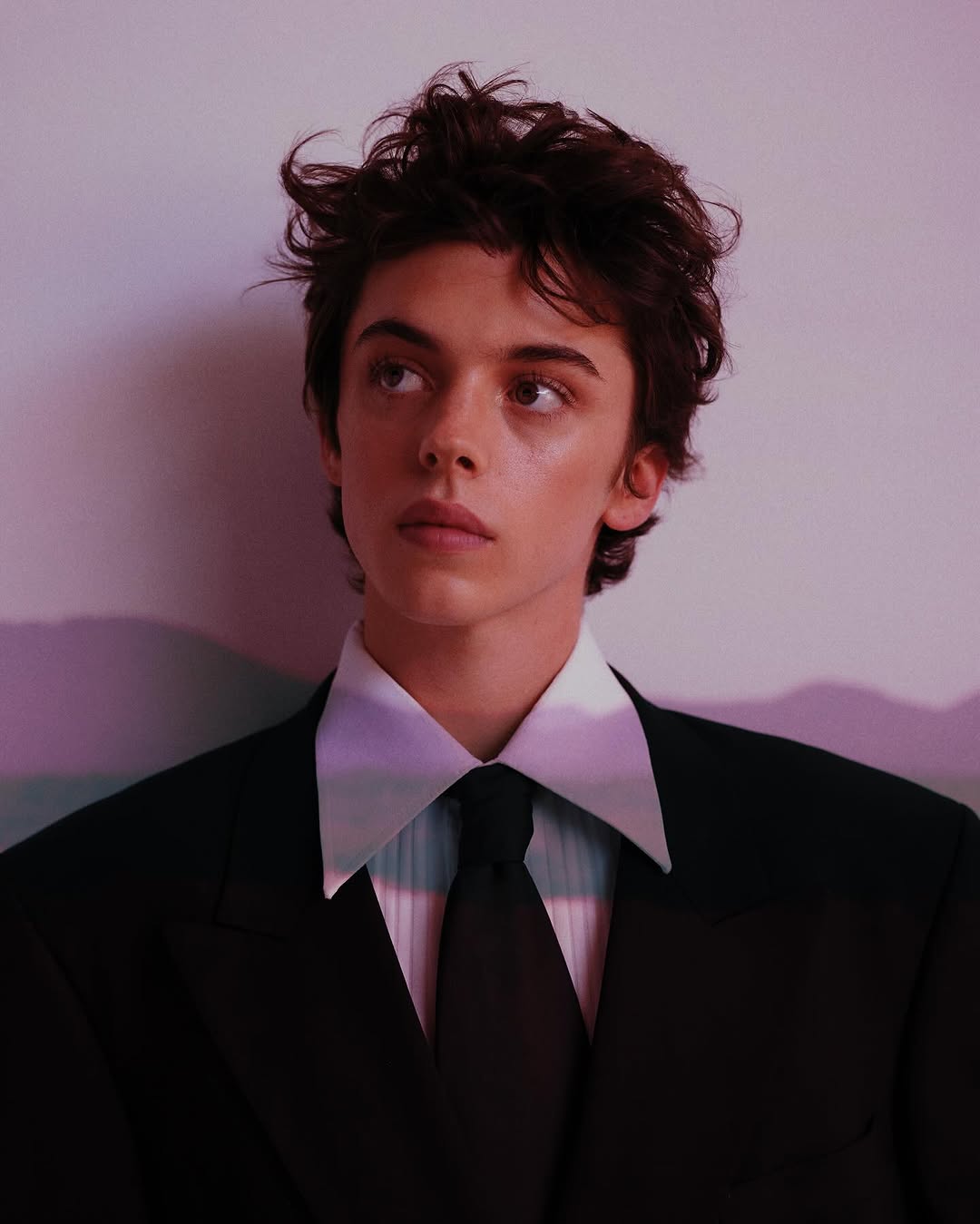 Portrait of a young adult with dark hair in a black suit and tie, gazing upwards, against a soft, pastel background with shadows.