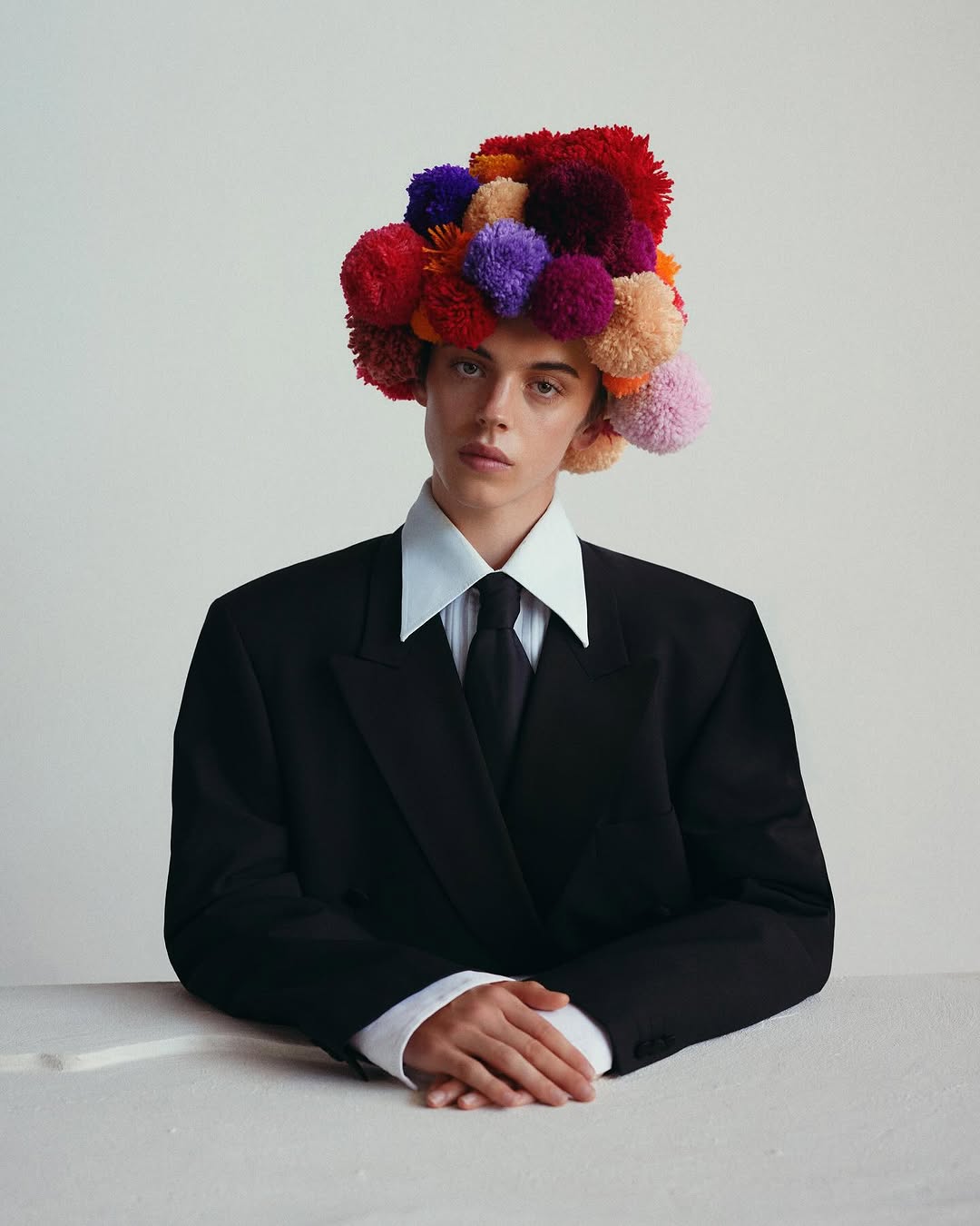 Young adult in suit with vibrant pompom hat, fashion portrait. Sharp collar, dark tie, hands clasped on light surface, neutral background, editorial.