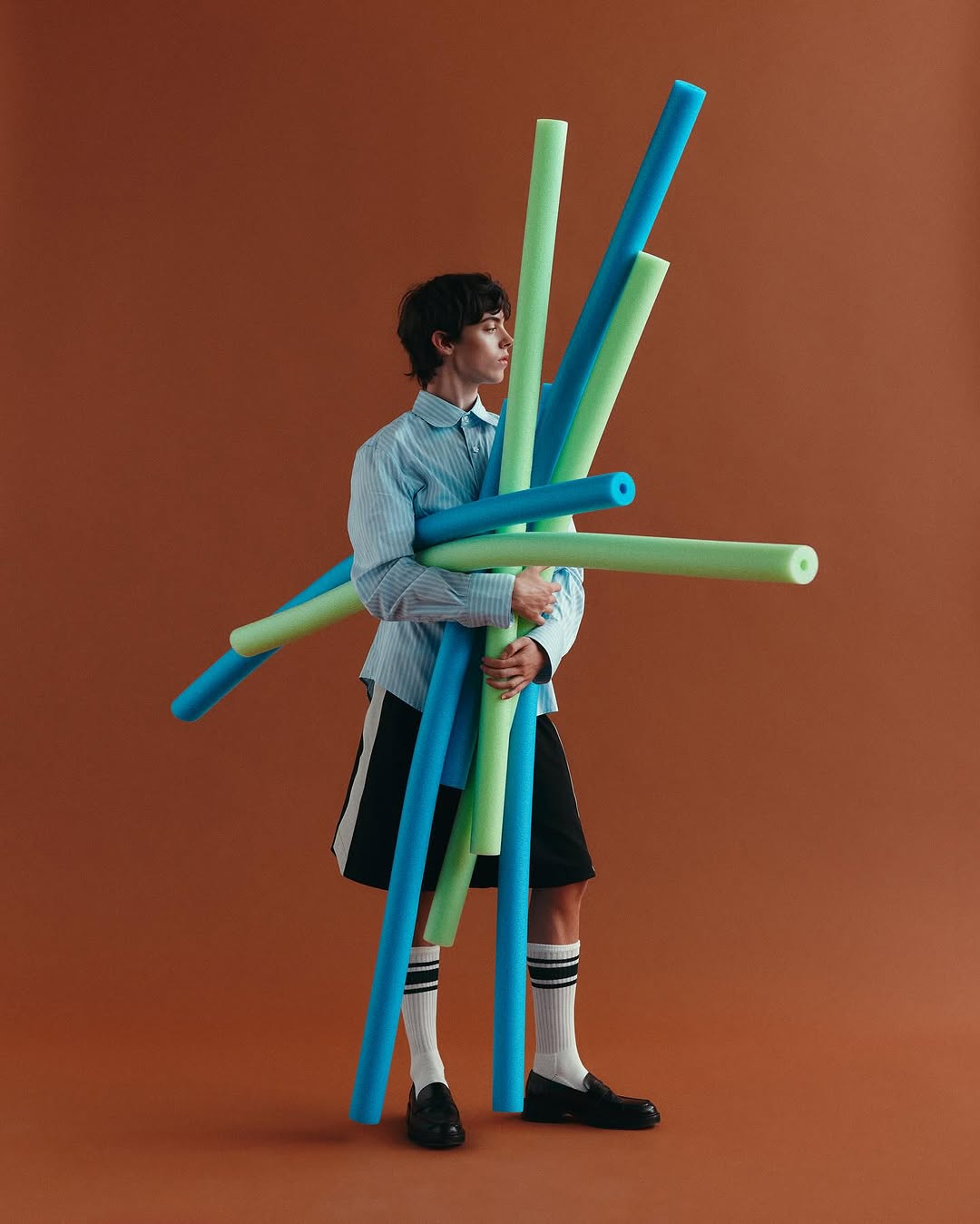 Person in striped shirt holds a bundle of blue and green pool noodles against an orange background in a studio setting.