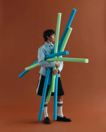 Person in striped shirt holds a bundle of blue and green pool noodles against an orange background in a studio setting.