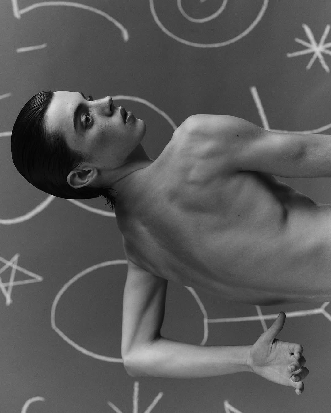 Monochromatic portrait of a shirtless young adult with slicked-back dark hair, posing against a grey background with white chalk drawings.