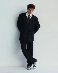 Young man in a black oversized suit and white shirt leaning against a plain white wall