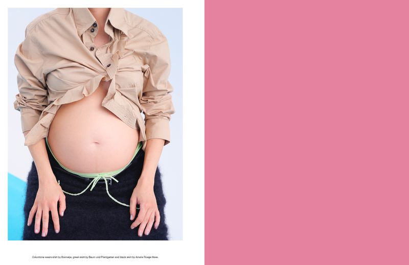 Close-up of a pregnant person in a partially buttoned tan shirt and black skirt with light green drawstrings, showcasing their bare stomach.