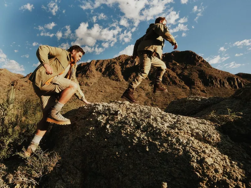 Two men wearing khaki outdoor jackets and hiking boots climbing and jumping across rocky mountain terrain under a blue sky.