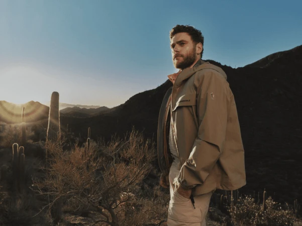 Model wearing a beige hooded performance jacket standing in a mountainous desert landscape with saguaro cacti at sunrise.