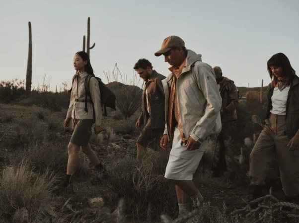 A group of models in neutral technical hiking jackets and shorts walking through a desert landscape with saguaro cacti.