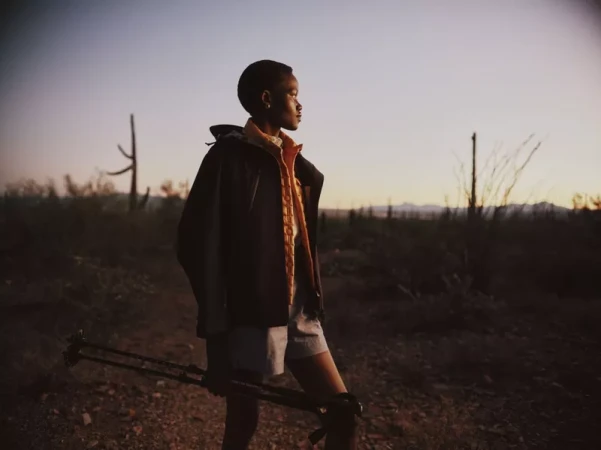 Model holding trekking poles in a desert landscape at dusk, wearing a dark outdoor jacket and an orange layered vest.