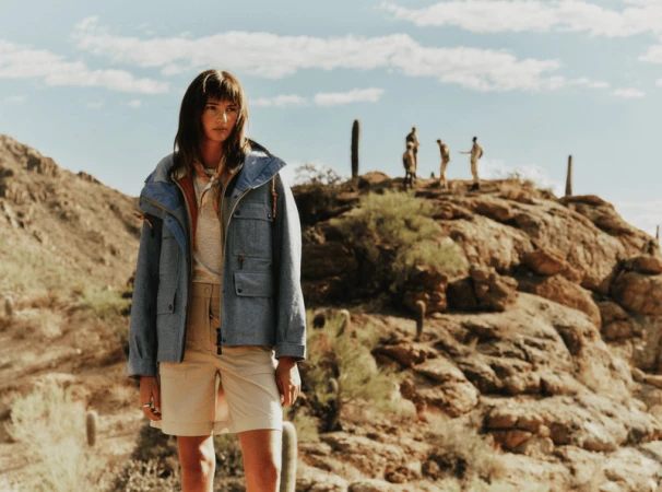 Model wearing a blue jacket and beige shorts standing in a desert landscape with rocky terrain and cacti under a blue sky.