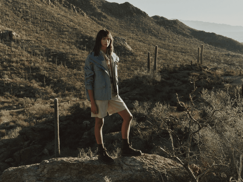 Model standing on a desert rock wearing a blue utility jacket, beige shorts, and hiking boots with saguaro cacti in the background.