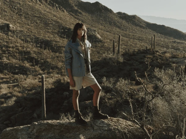 Model standing on a desert rock wearing a blue utility jacket, beige shorts, and hiking boots with saguaro cacti in the background.