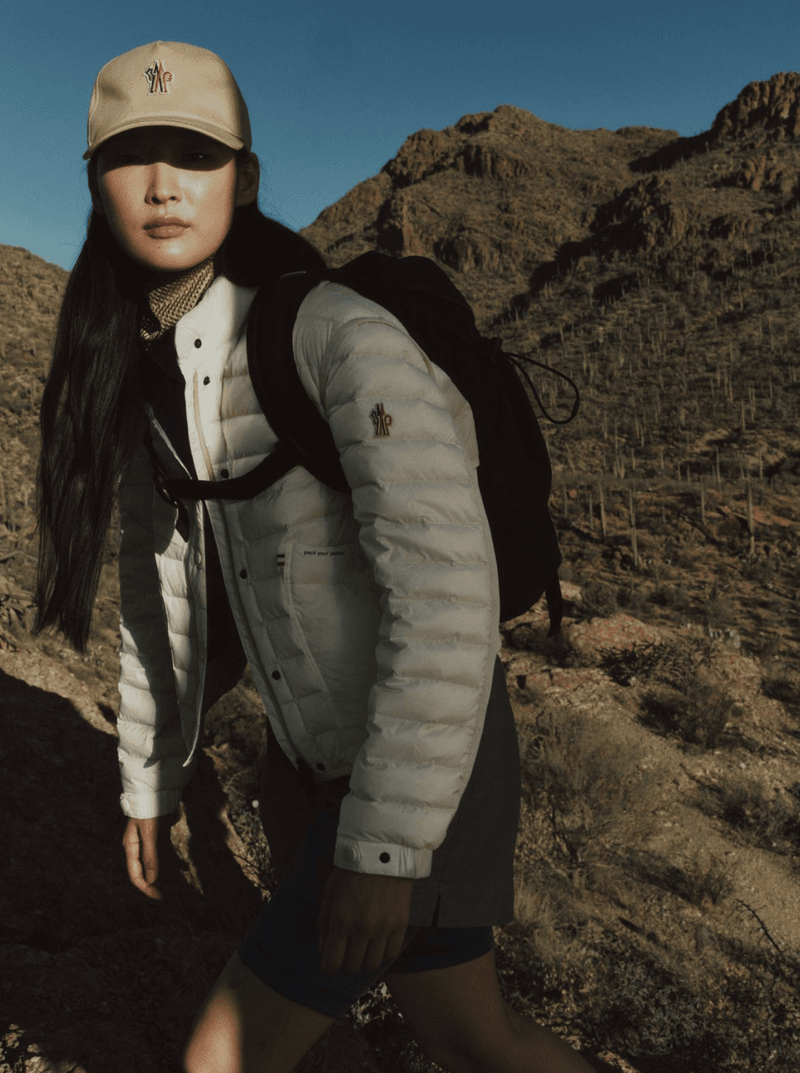 East Asian woman wearing a cream puffer jacket, beige baseball cap, and black backpack hiking along a rocky mountain ridge.
