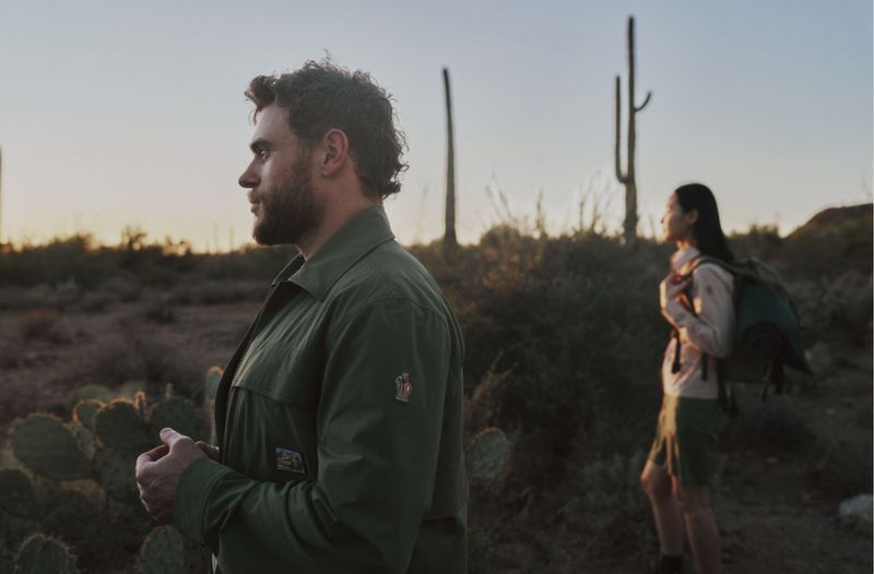 Man in green jacket in profile with a woman standing among desert cacti at sunset for Moncler Grenoble.