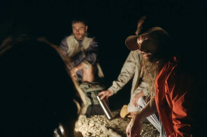 A man in a wide-brim hat and bright orange jacket crouching near a campfire at night with companions on rocky terrain.
