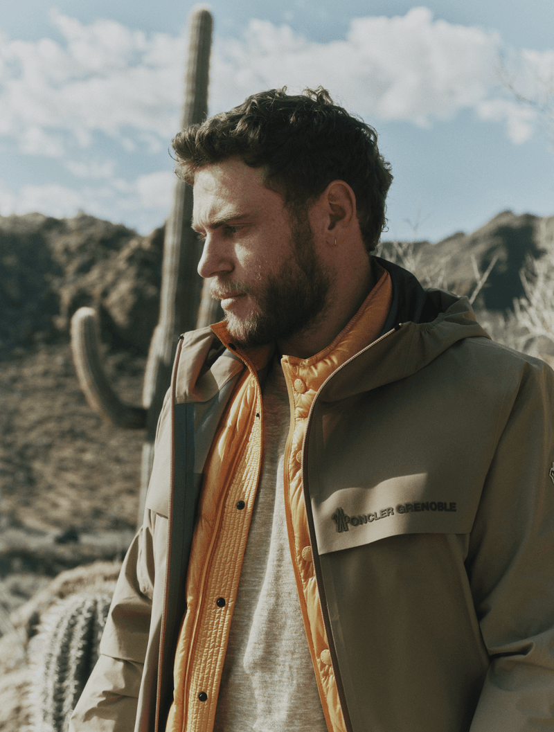 Man with beard wearing a Moncler Grenoble beige hooded jacket and yellow quilted vest outdoors in a desert with cacti.