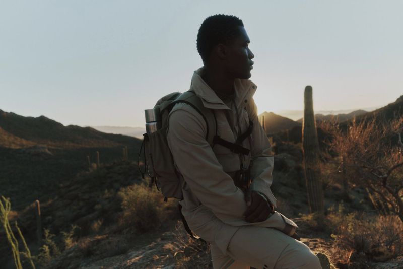 Black man wearing utility jacket with backpack standing outdoors in mountainous desert landscape at sunset