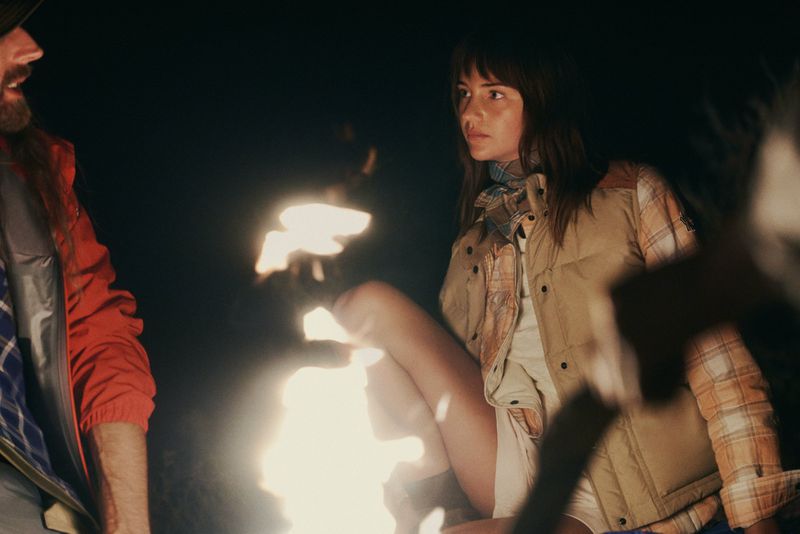 Mia Regan wearing a beige puffer vest and plaid shirt seated by a glowing campfire at night beside a man in a red jacket.