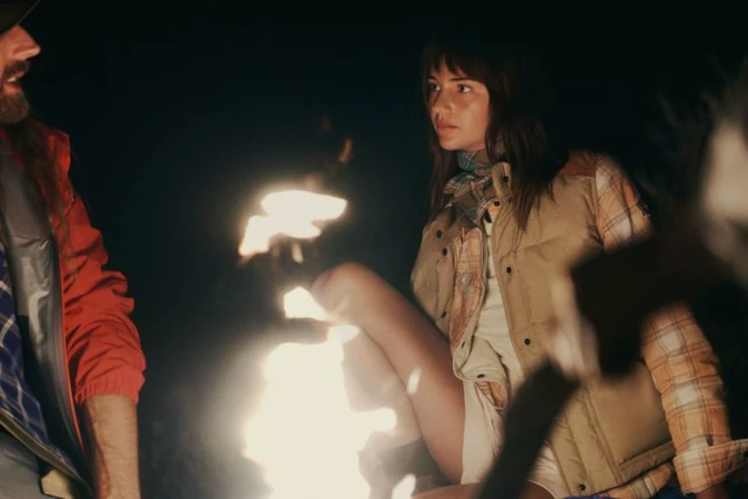 Mia Regan wearing a beige puffer vest and plaid shirt seated by a glowing campfire at night beside a man in a red jacket.