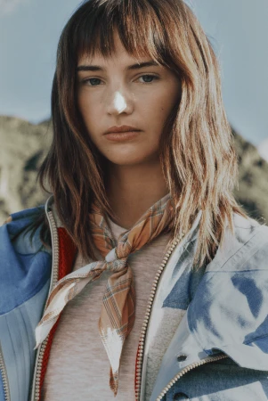 Model Mia Regan looking at camera wearing a blue patterned outdoor jacket and plaid neck scarf against a mountain backdrop.